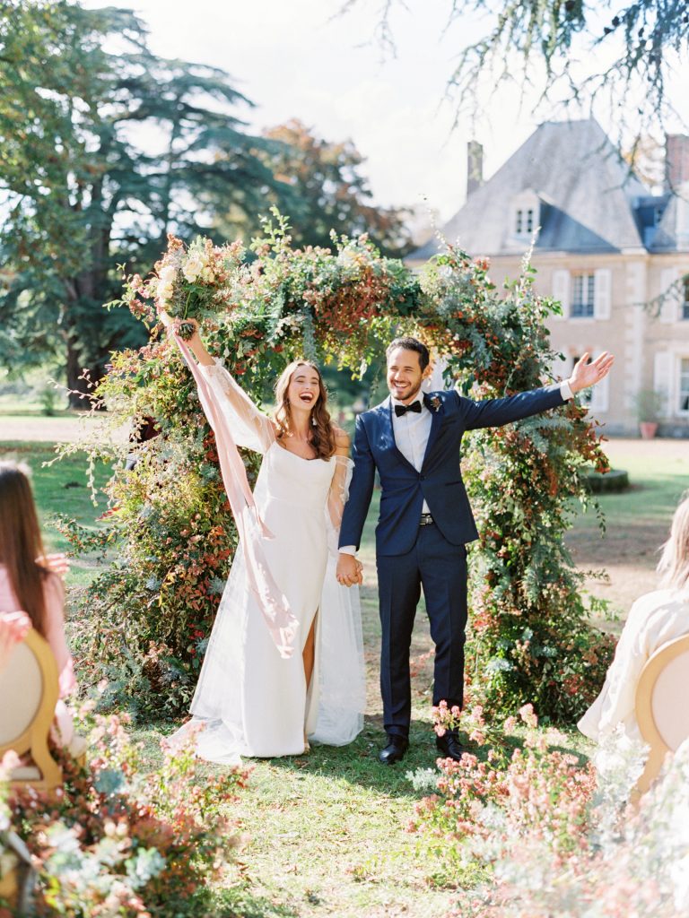 bride and groom at ceremony in paris france. happy bride and groom. bride is wearing a white couture wedding gown. groom has on a tuxedo from the black tux. bride is holding bouquet in the air. guests are clapping.