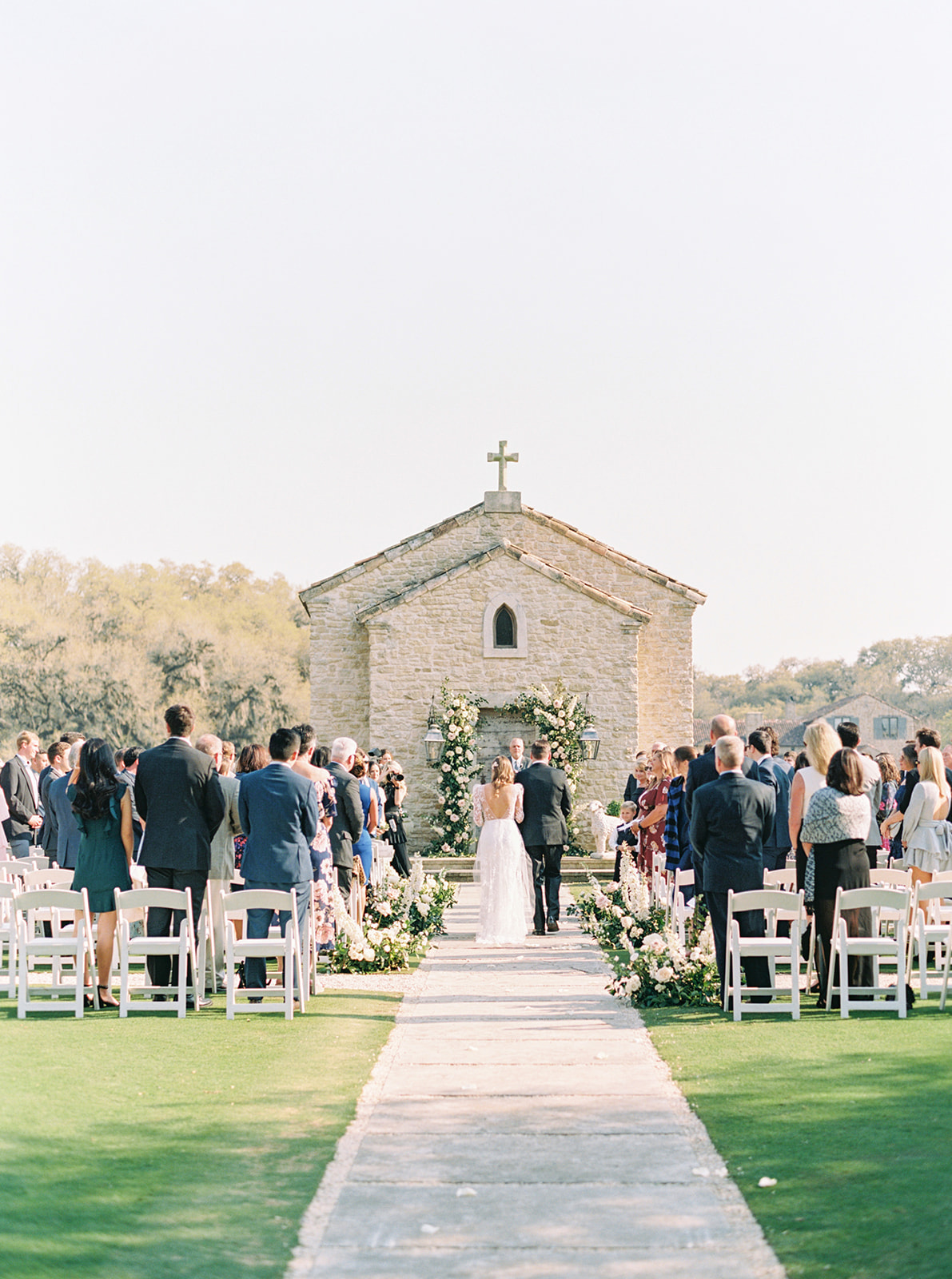 Bride and groom standing at the altar by the end of the aisle along with their wedding guests during their outdoor romantic chapel wedding
