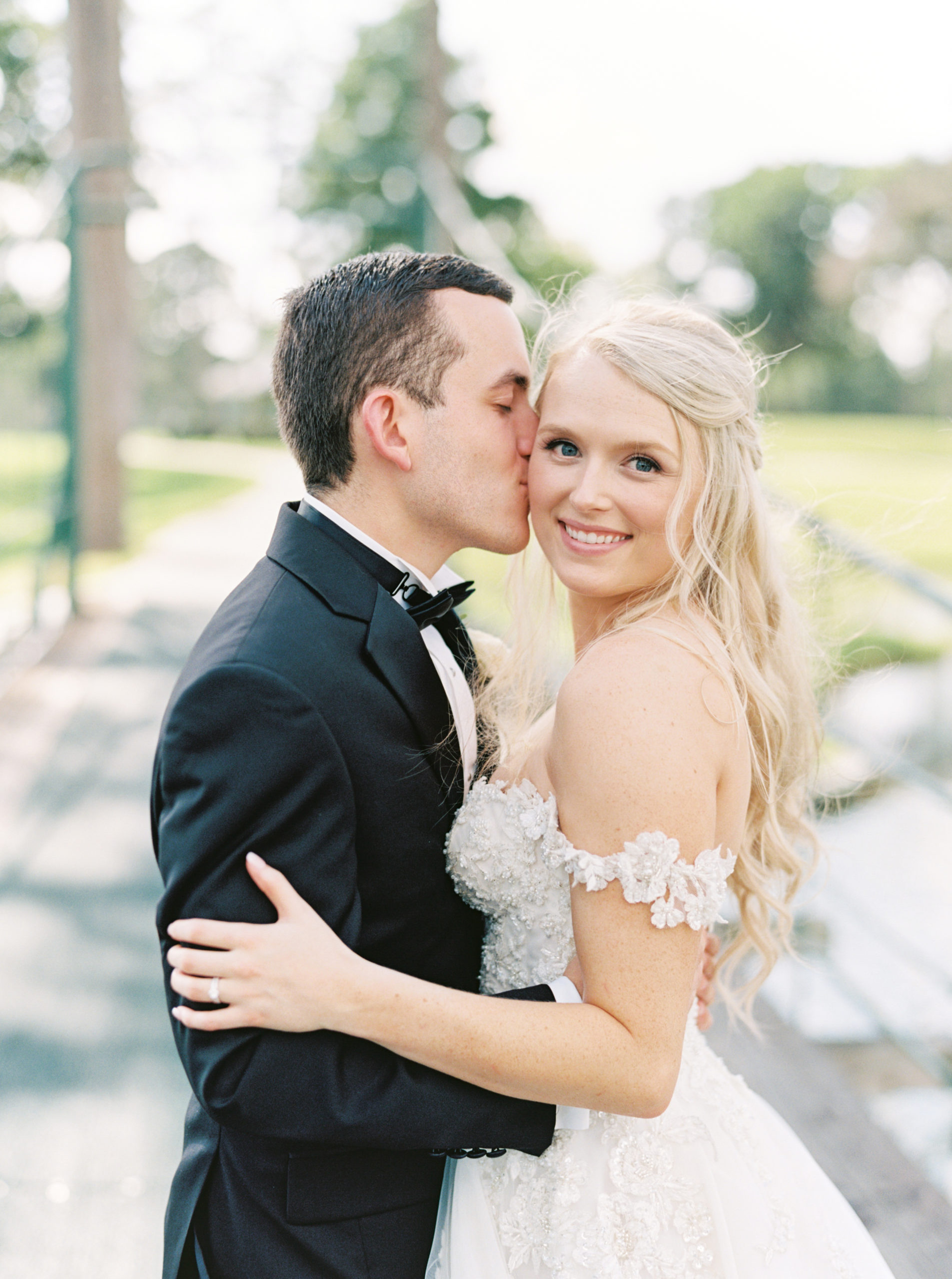 Bride looks at the camera enchantingly as the groom gives her a kiss on the cheek during their wedding shoot with Mackenzie Reiter